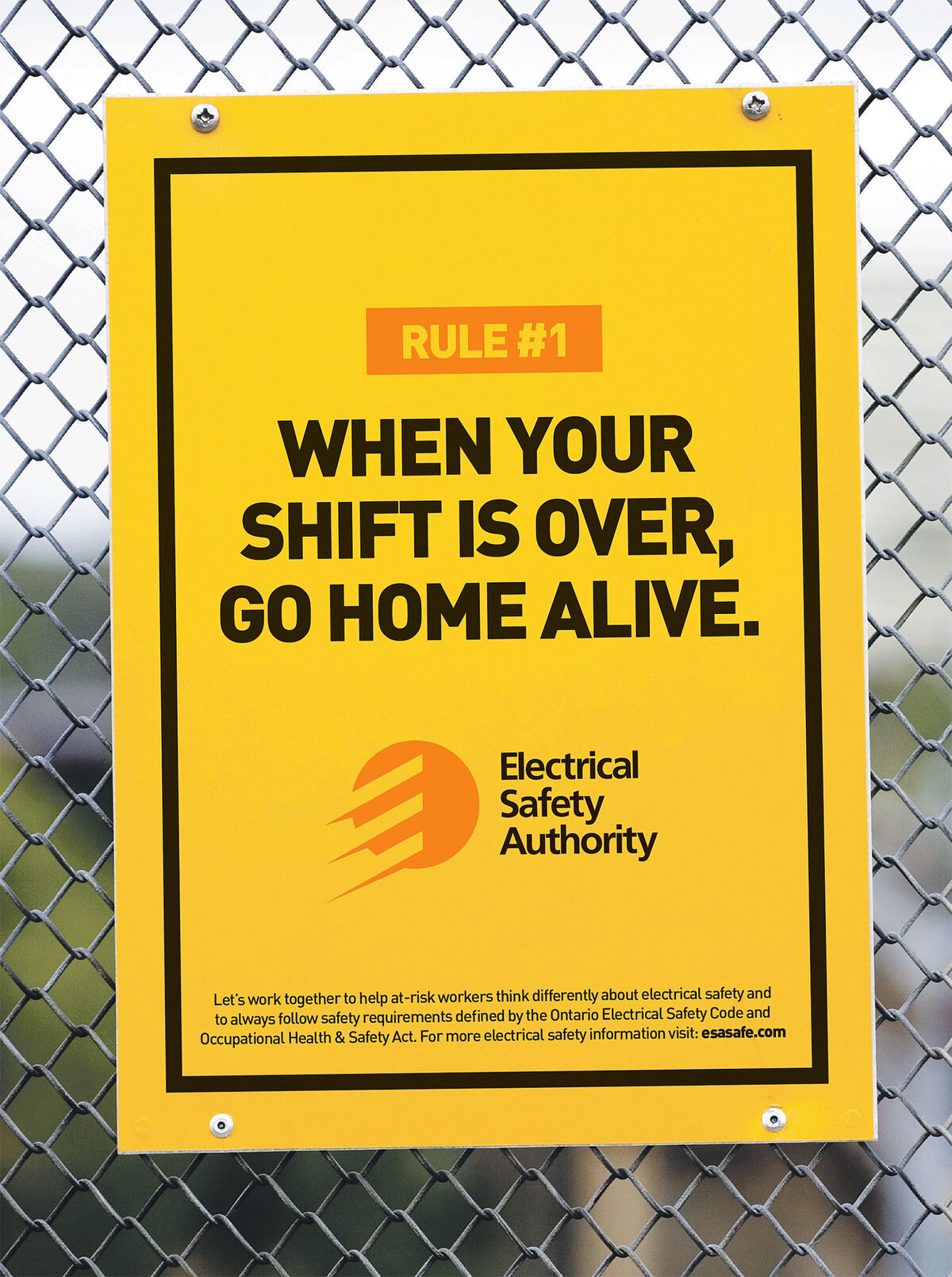 An Electrical Safety Authority trade ad reading 'When your shift is over, go home alive,' posted on a chain-link fence at a work site