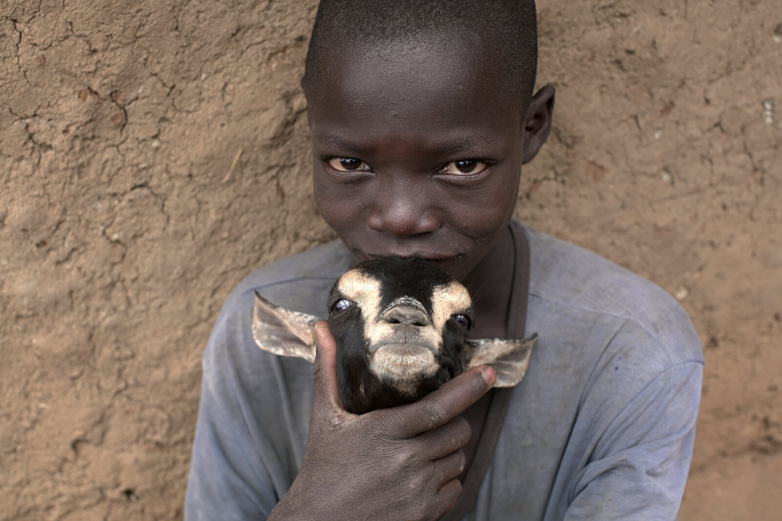 A young boy holds a goat up to his face against a mud wall in rural Uganda
