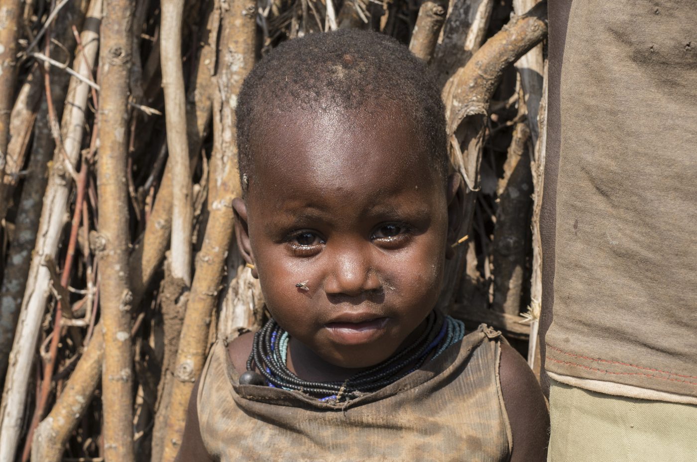 Lochap&rsquo;s young little sister in a worn dress, looking at the camera before a stick fence